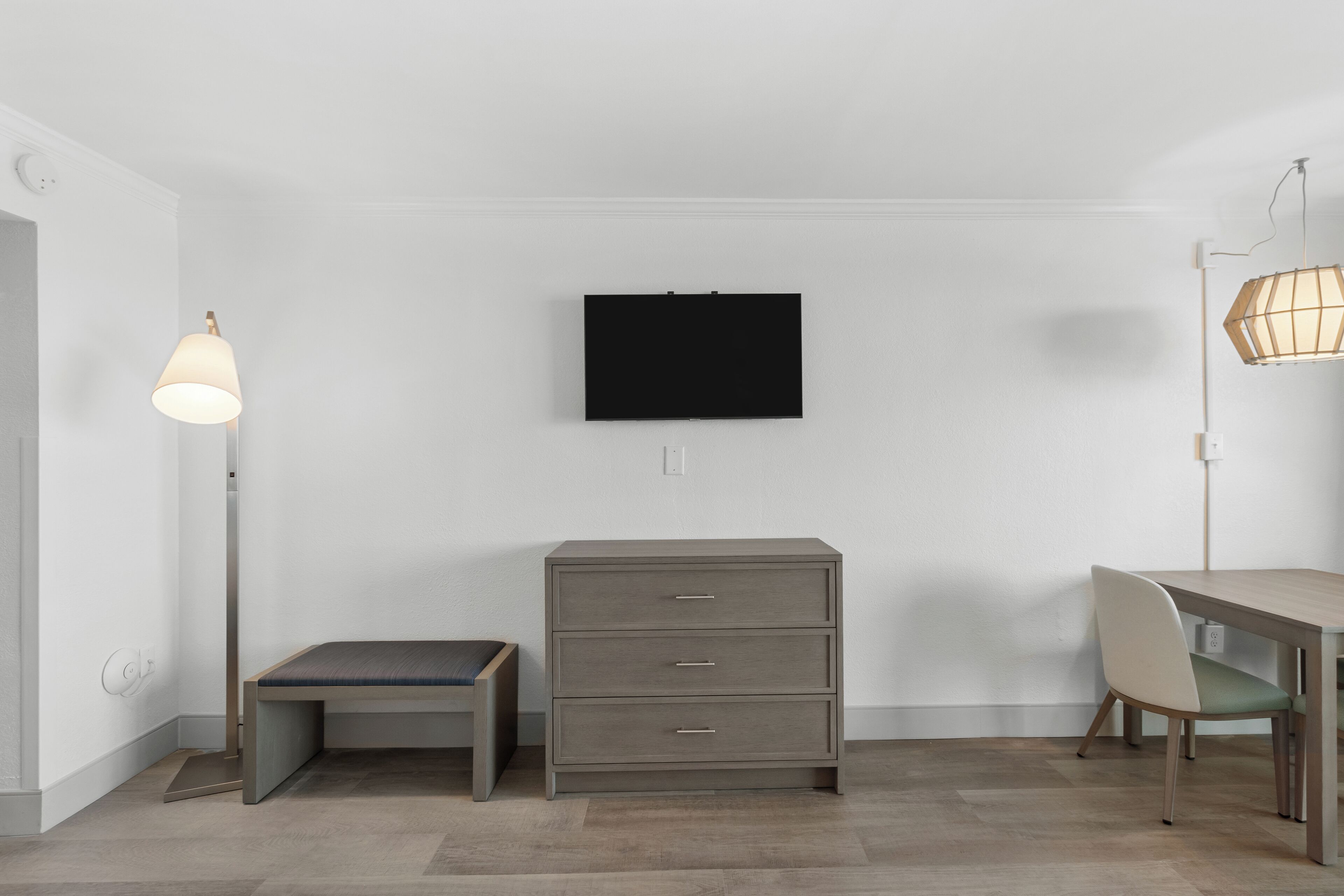 Una habitación con televisión de pantalla plana montada en la pared, un aparador gris, un banco, una mesa con una silla y una lámpara de pie.