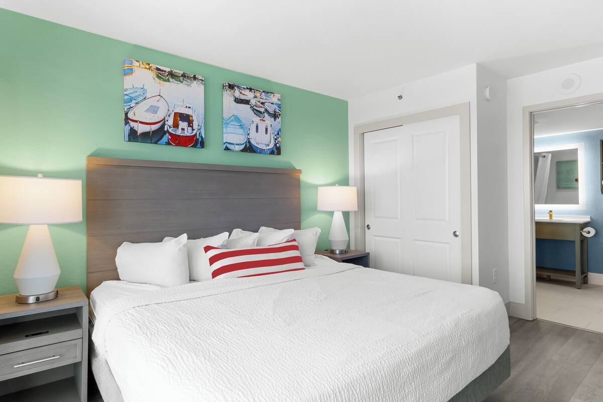 A hotel room with a large bed, two bedside tables with lamps, a headboard, and two wall-mounted artworks depicting boats.