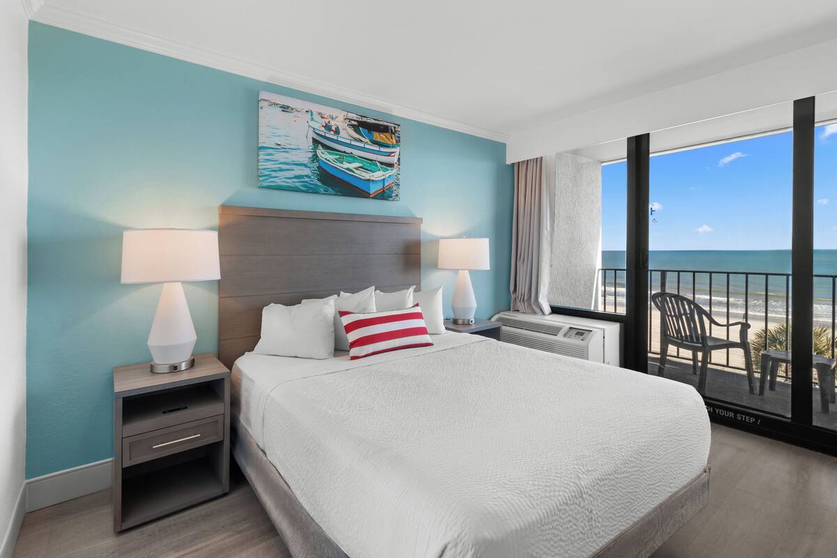 A hotel room with a large bed, two bedside tables with lamps, a painting of boats on the wall, and a view of the beach through a large window.
