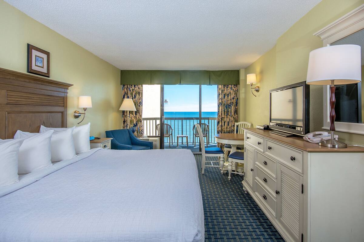 A hotel room with a large bed, a desk with a chair, a television, and a view of the ocean.