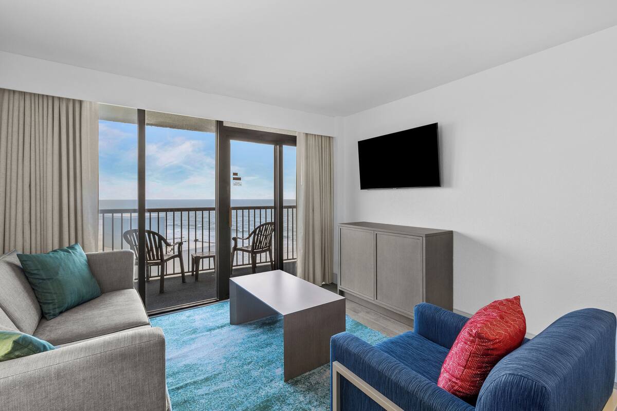 A hotel room with a balcony, a TV, a sofa, a chair, and a coffee table.