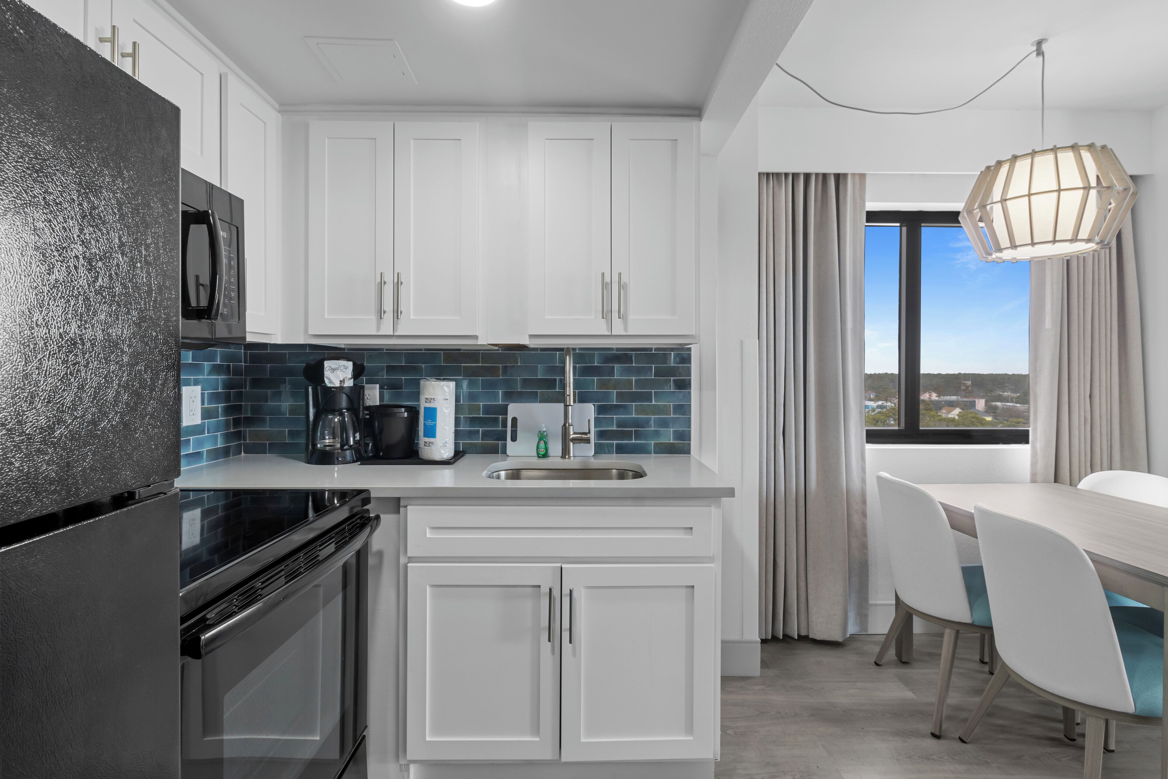 A modern kitchen with white cabinets, a black refrigerator, a blue-tiled backsplash, and a dining area with a table and chairs.