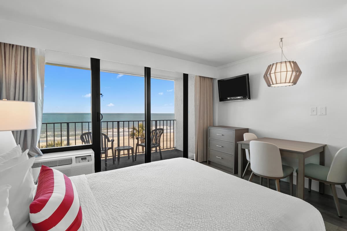 A hotel room with a large bed, a desk, a chair, a television, and a view of the beach.