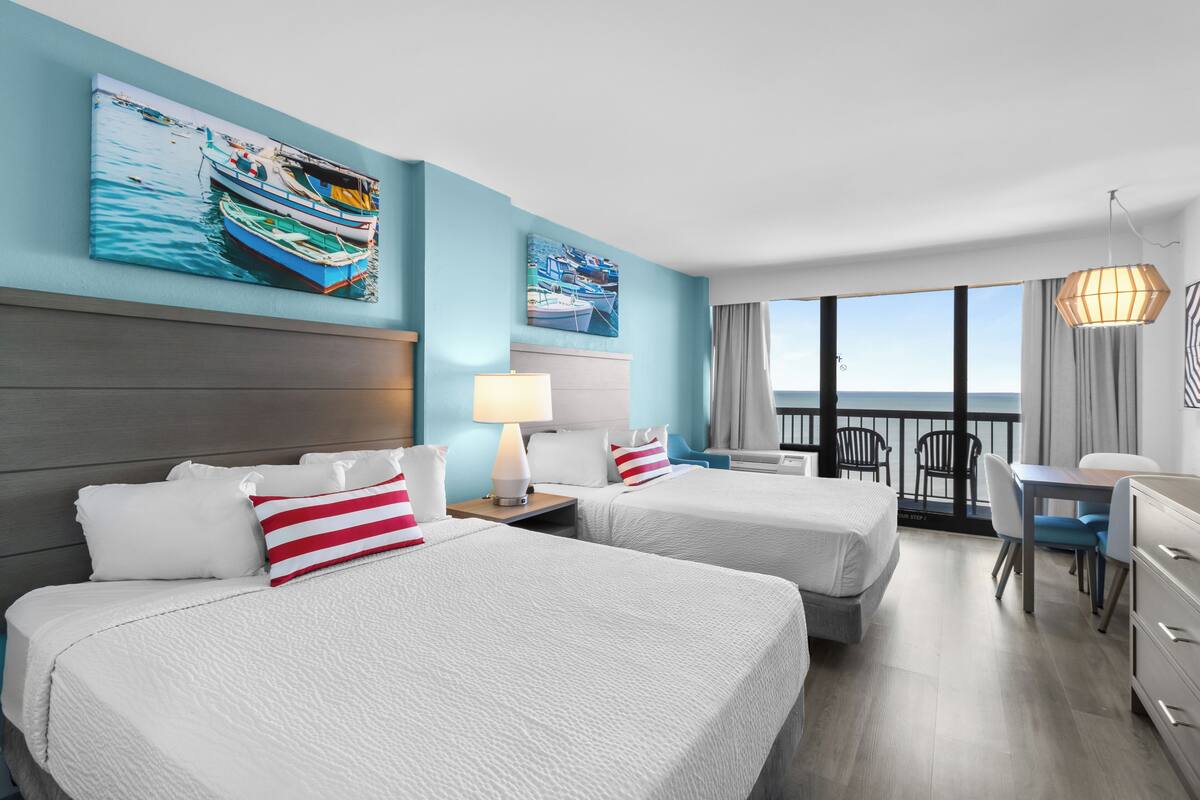 A hotel room with two beds, a desk, and a balcony with a view of the sea.