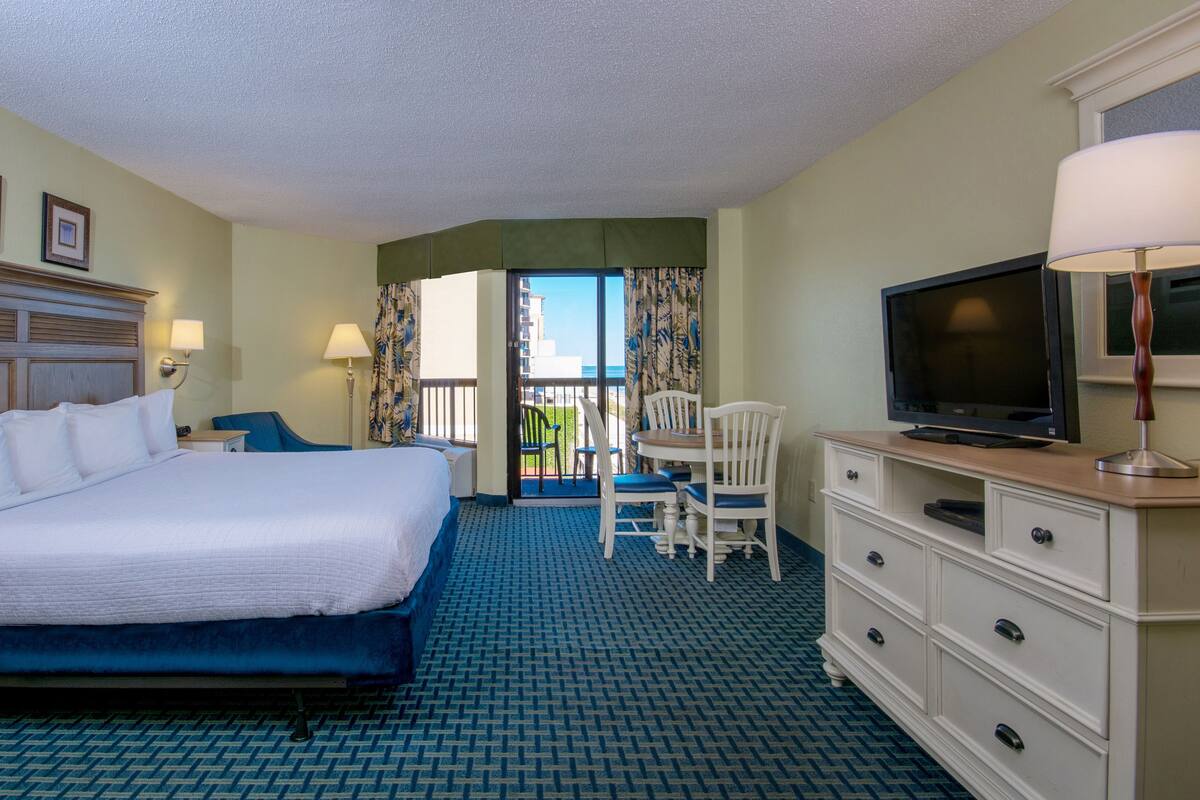 A hotel room with a large bed, a desk with a chair, a television, and a view of the ocean through the balcony doors.