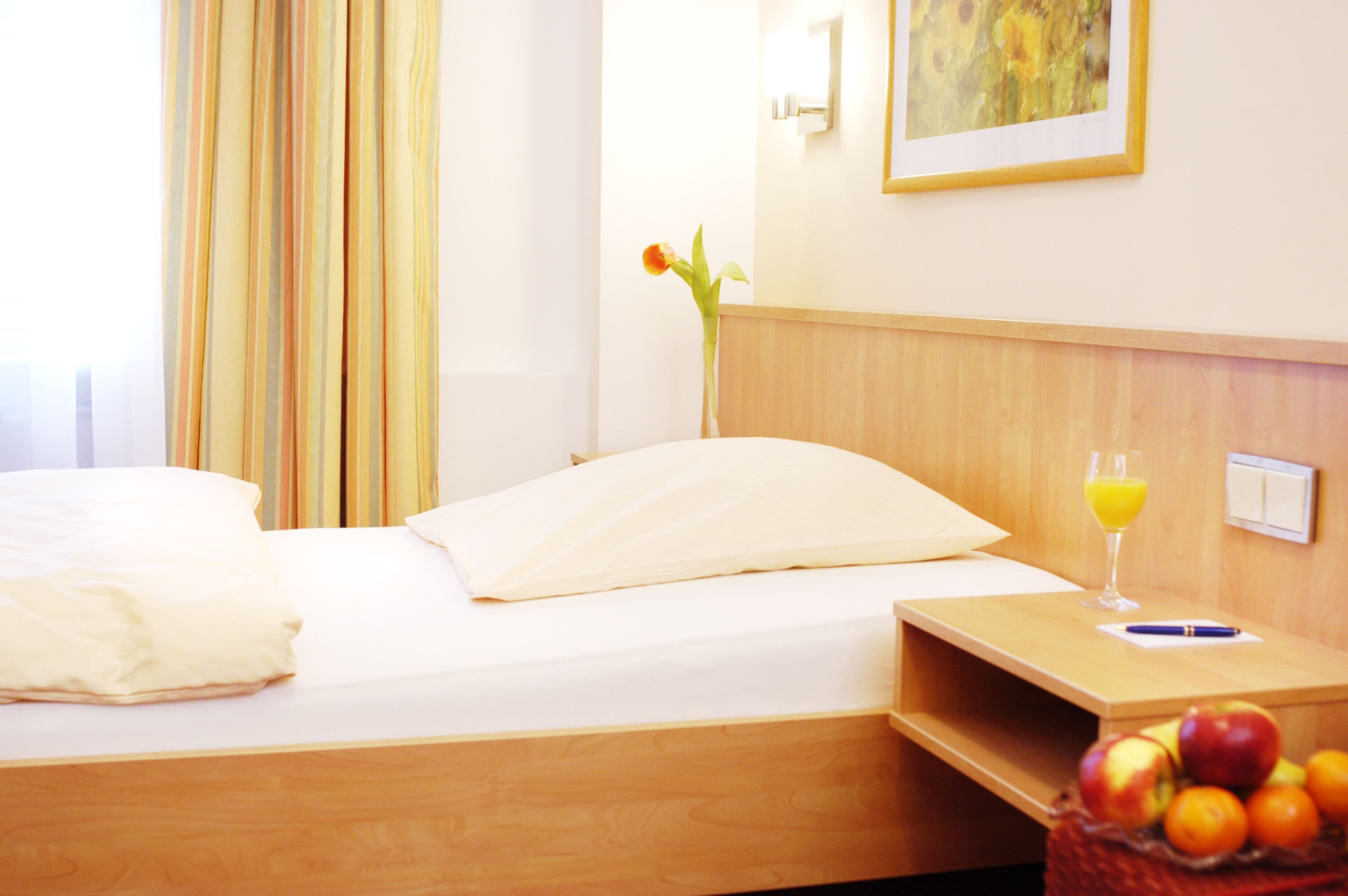 A hotel room with a bed, a bedside table with a glass of orange juice, a vase with a flower, and a basket of fruit.