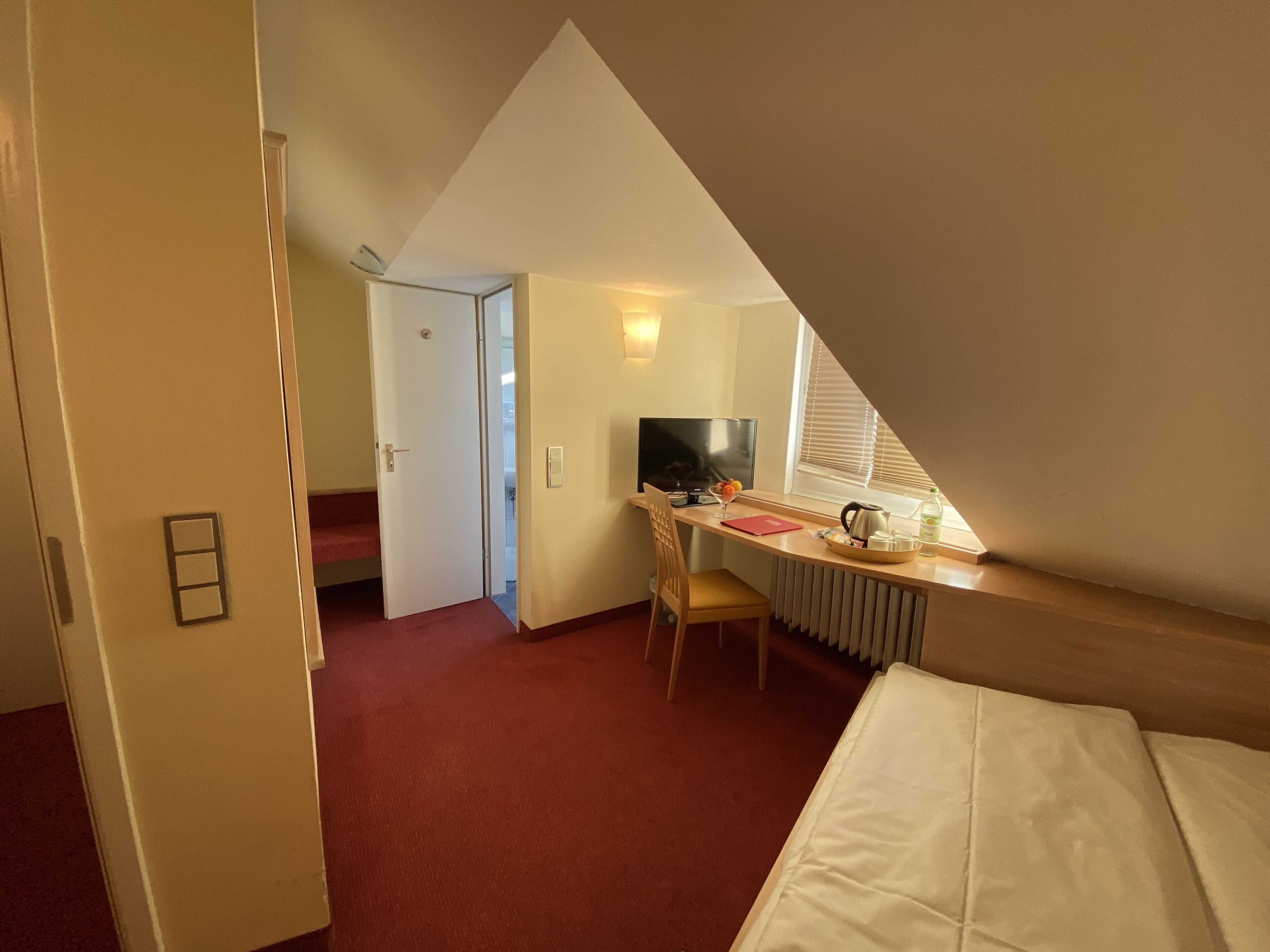Ein Schlafzimmer mit schrägen Wänden, einem Bett, einem Schreibtisch mit Stuhl, einem Fernseher und einer Heizung.