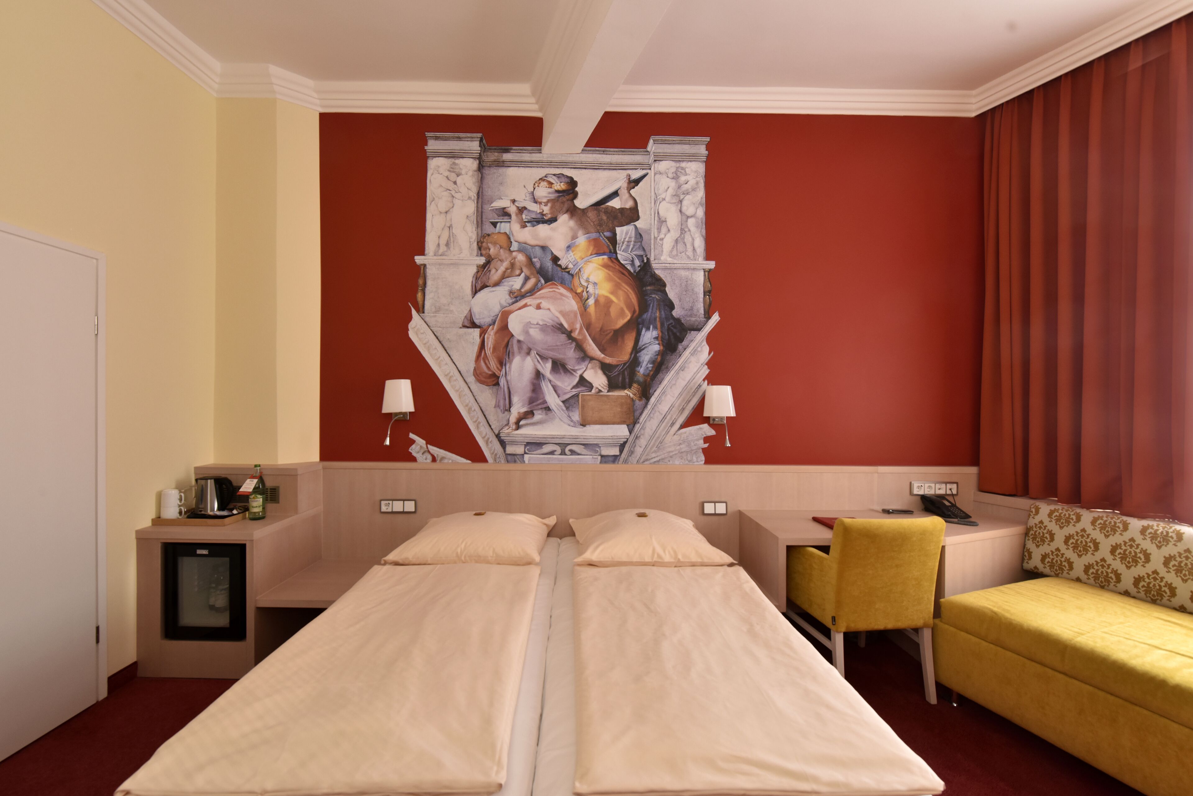 Ein Hotelzimmer mit zwei Betten, einem Schreibtisch, einem Stuhl, einem kleinen Tisch und einem Wandgemälde.