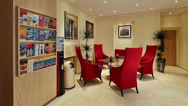 A modern hotel lobby with red chairs, a coffee machine, and a wall with travel brochures. - Hotel ADRIA München (Munich)