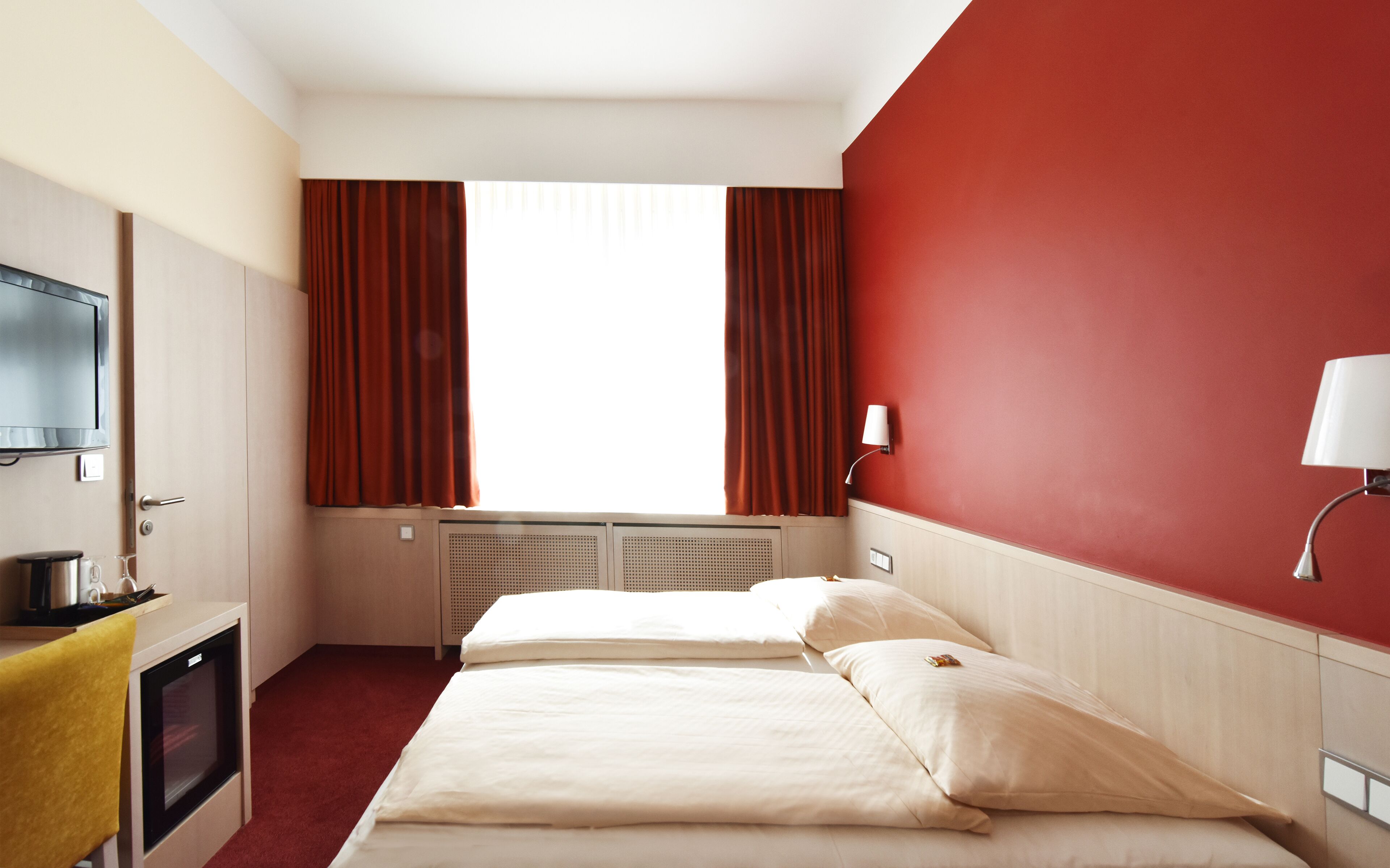 Ein Hotelzimmer mit zwei Betten, einem Fernseher, einem Schreibtisch mit Wasserkocher und einer roten Wand.
