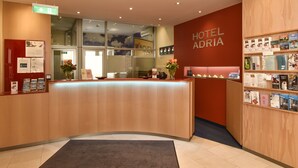 A hotel reception desk with a wooden counter, a red wall with the hotel name, and informational brochures on the right. - Hotel ADRIA München (Munich)