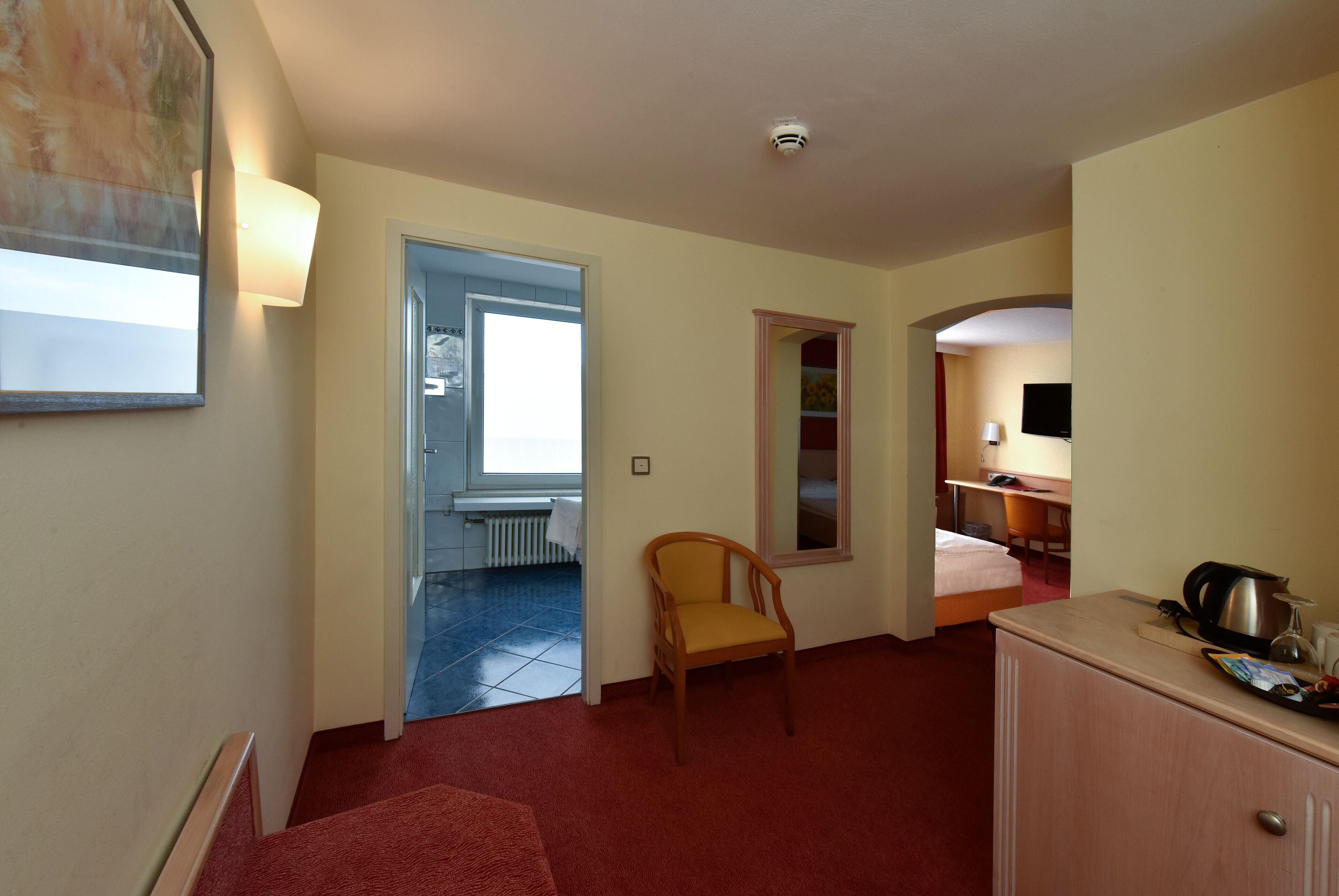 A hotel room with a red carpet, a wooden chair, a bed, a desk with a chair, a television, and a kitchenette area.