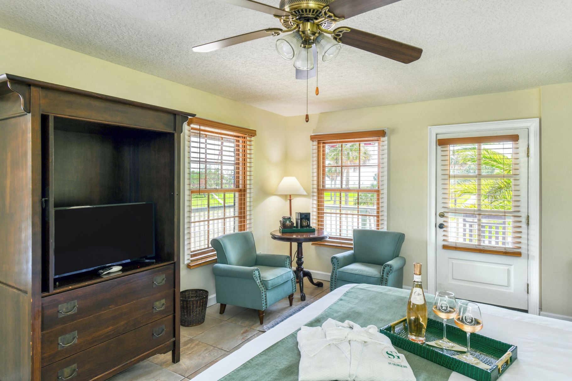 A hotel room with a television, two armchairs, a table with a bottle and glasses, and a bed with a green bedspread.