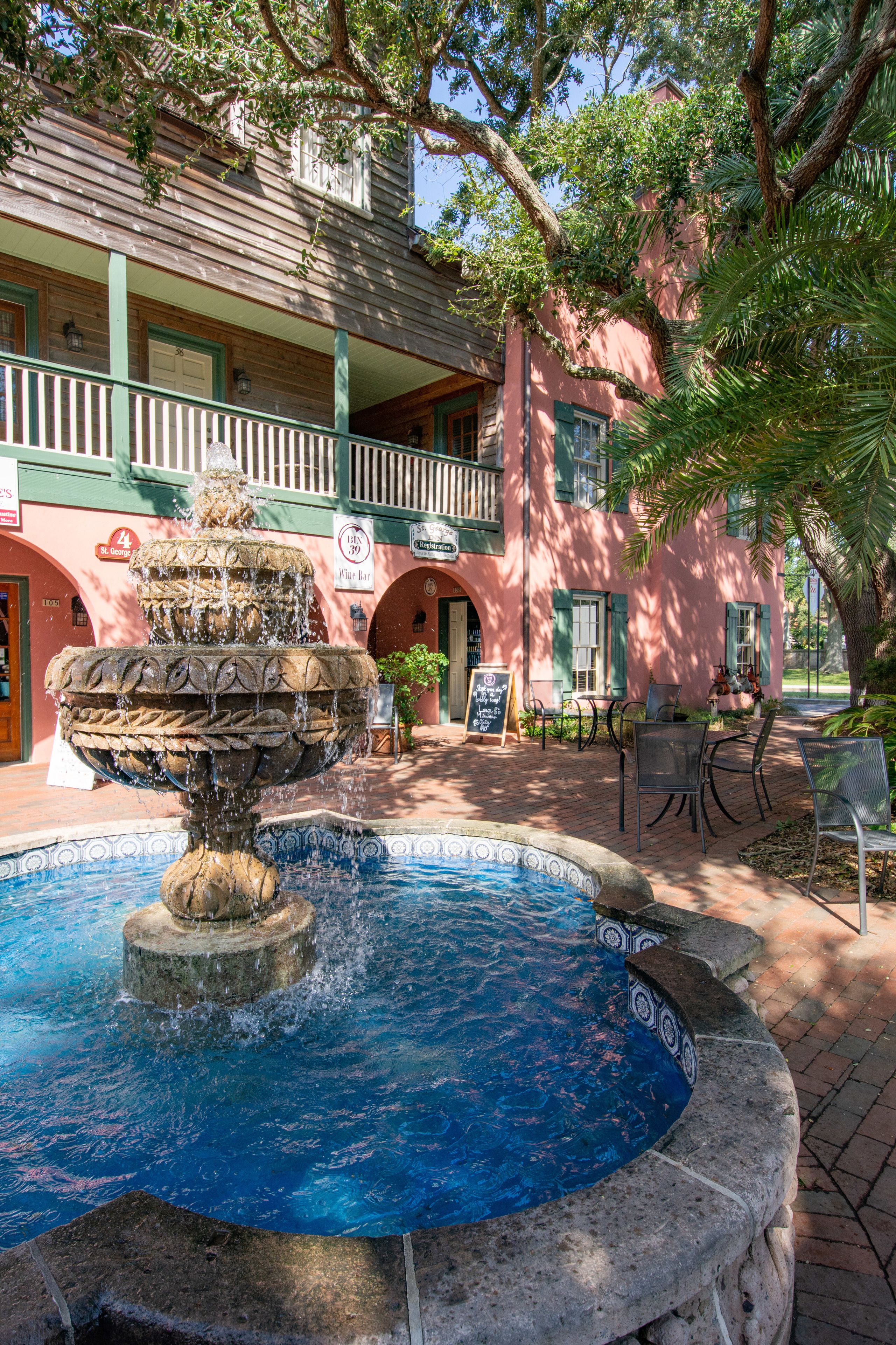 A courtyard with a stone fountain, outdoor seating, and a building with a balcony.
