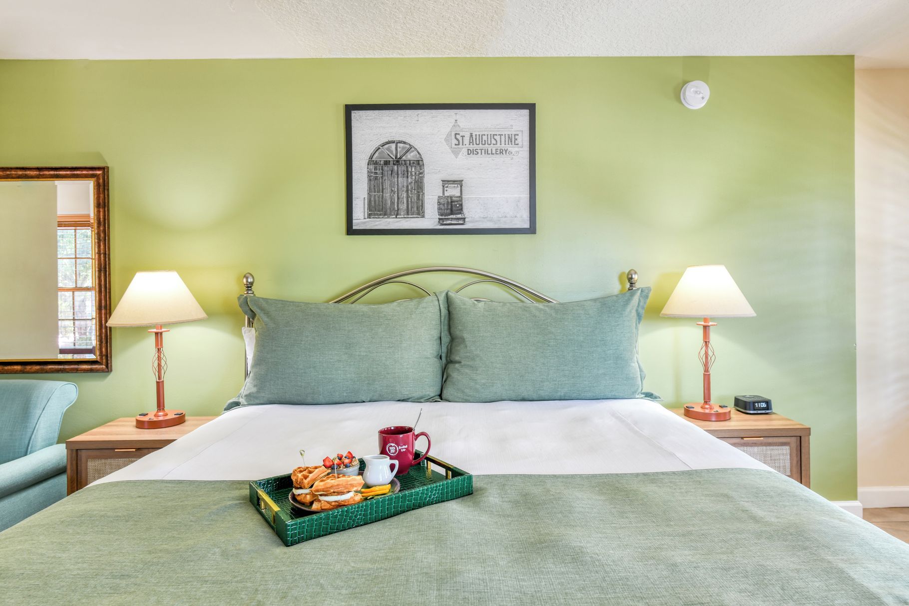 Una habitació d'hotel amb un llit, dues làmpades de nit, un quadre al mur i una safata amb guaïlles i una tassa damunt el llit.