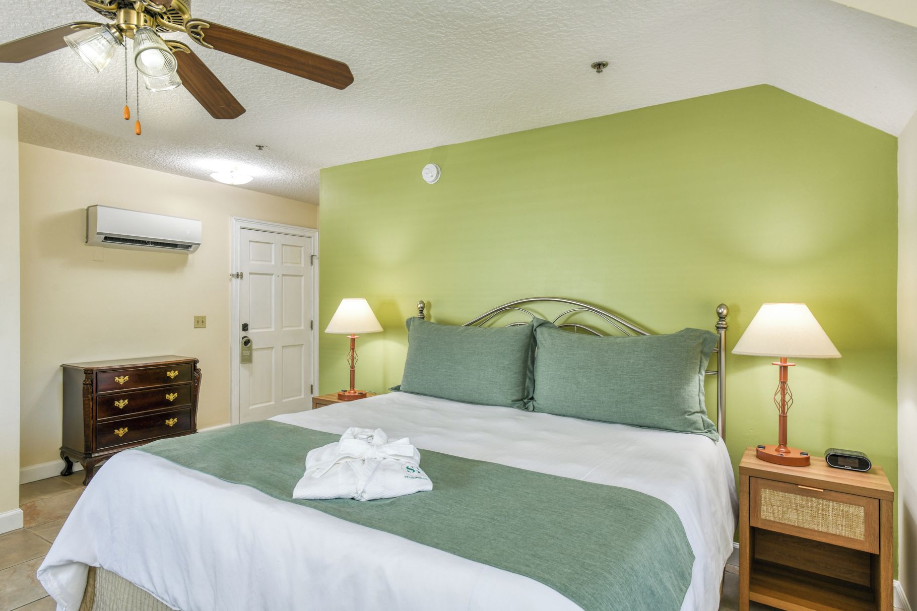 Een hotelkamer met een bed, twee nachtkastjes met lampen, een plafondventilator, een airconditioner, een houten dressoir en een groene wand.