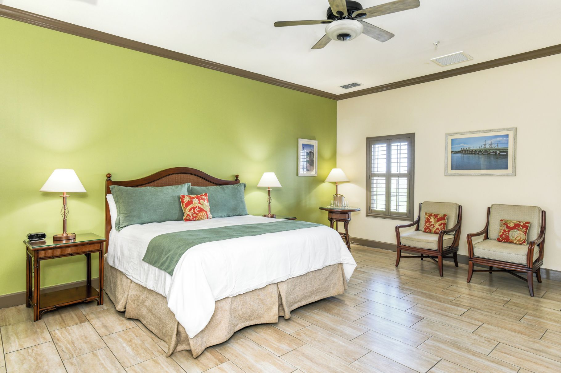 A hotel room with a bed, two bedside tables with lamps, a ceiling fan, two chairs, and a window with a view.