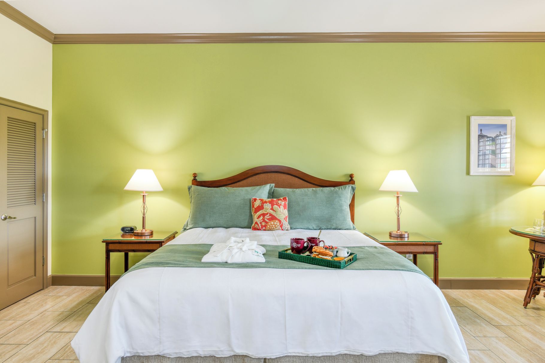 A hotel room with a bed, two bedside tables, a nightstand, a tray with breakfast items, and a framed picture on the wall.