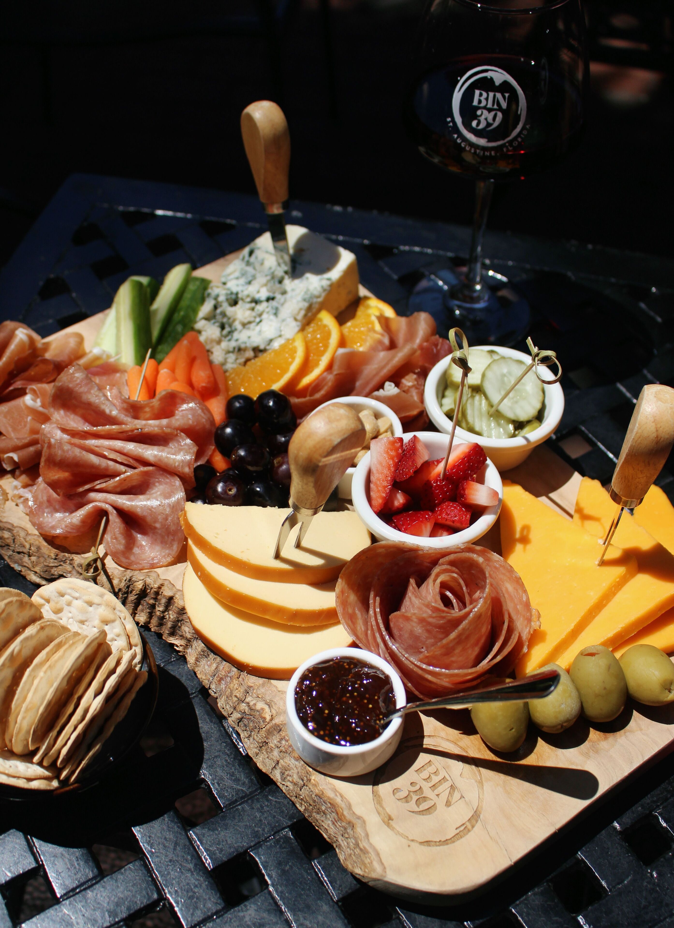Un tagliere con salumi, formaggi, frutta e un bicchiere di vino su un tavolo in metallo.