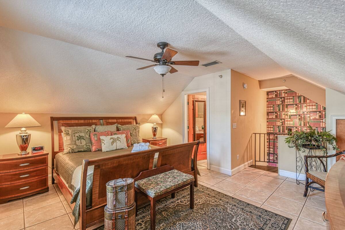 A bedroom with a bed, bedside tables, a chair, a ceiling fan, a bookshelf, and a staircase.