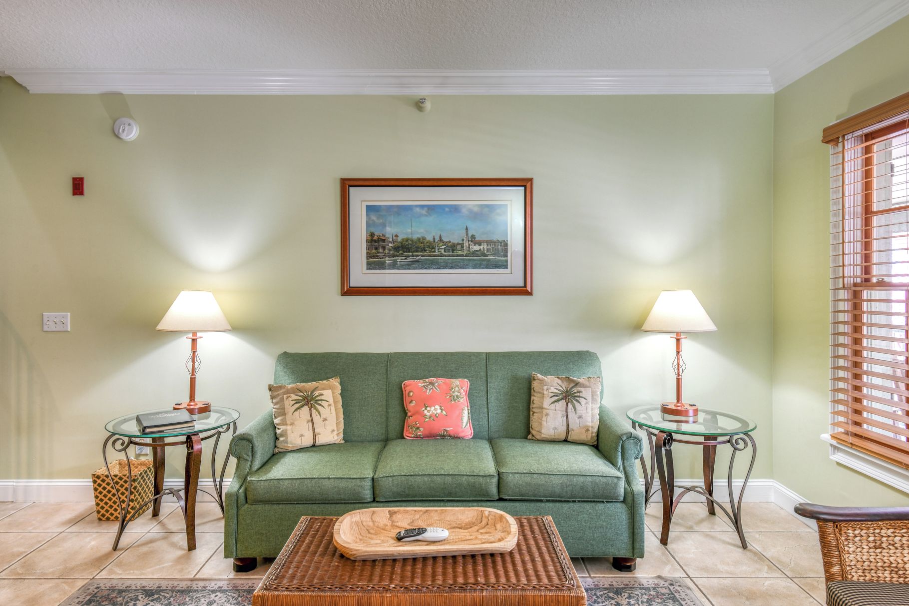 A living room with a green sofa, two side tables with lamps, a framed picture on the wall, and a window with blinds.