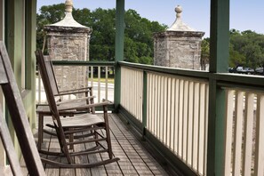 A wooden rocking chair on a balcony with a green railing and stone pillars.