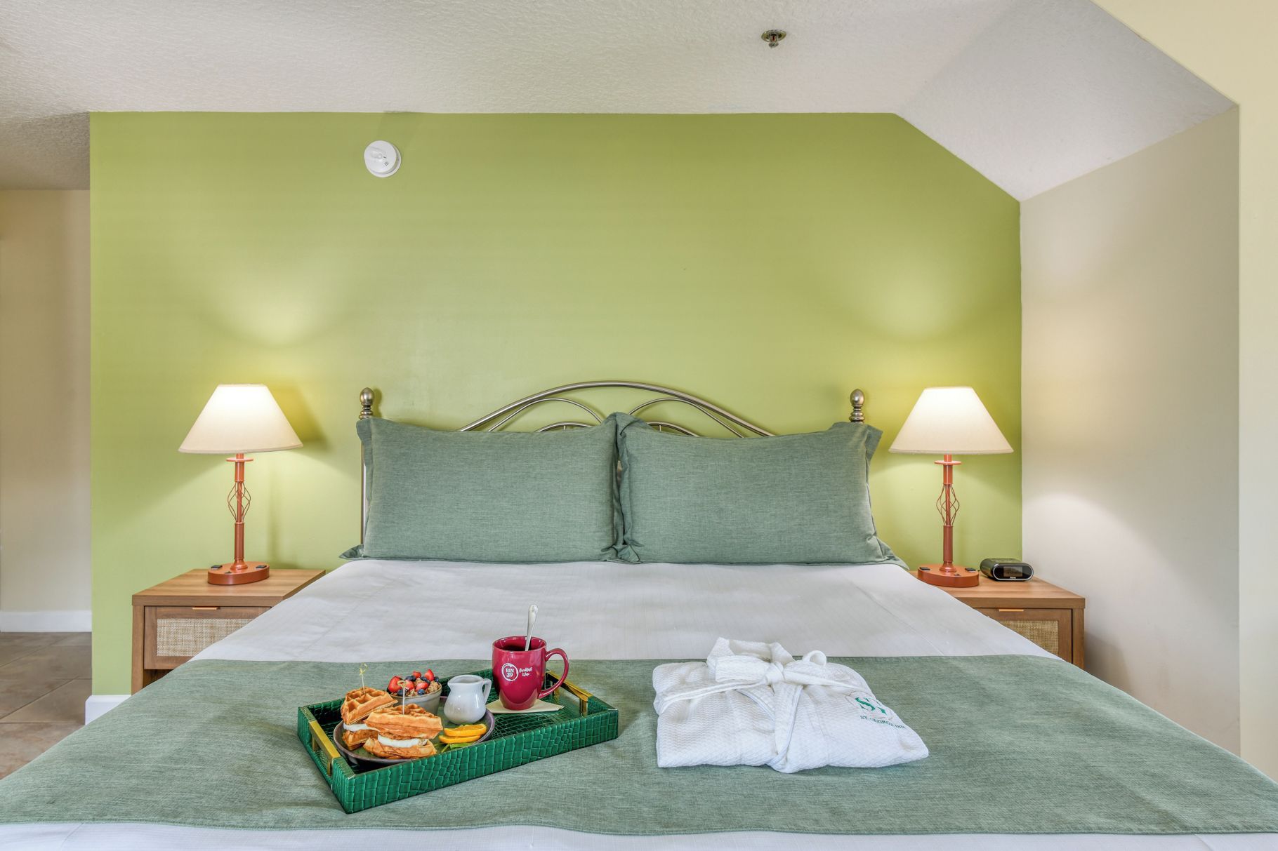 A hotel room with a bed, two bedside lamps, a tray with waffles and a mug, and a robe on the bed.