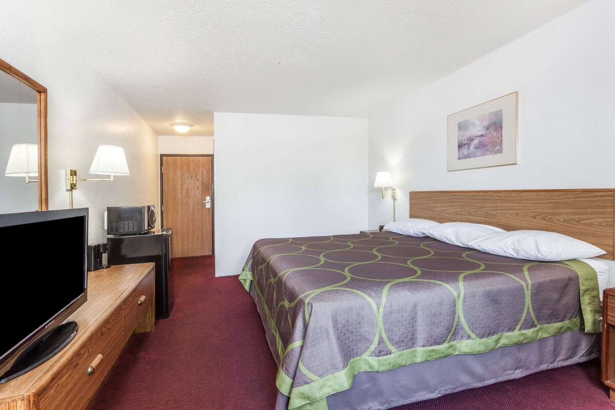 Habitación de hotel con cama, televisor sobre una cómoda de madera, escritorio con lámpara y un cuadro en la pared.