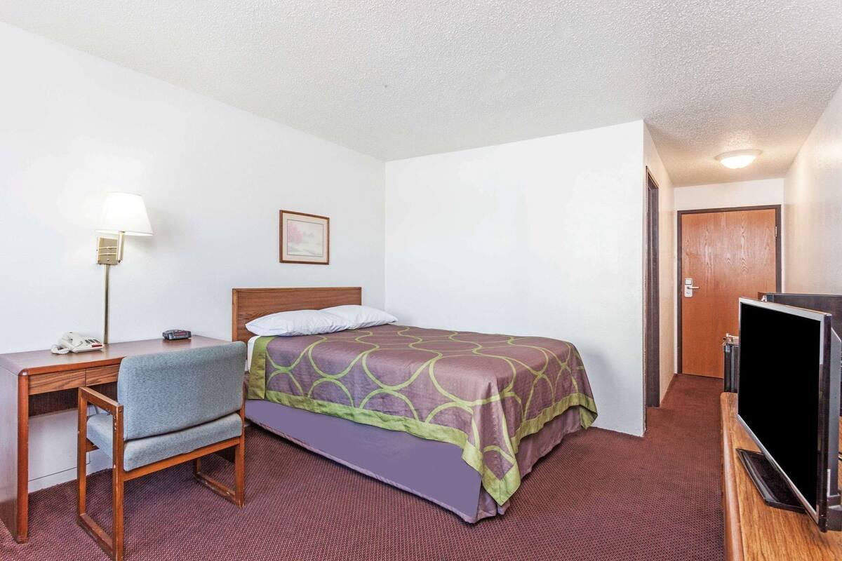 Una habitación de hotel con cama, escritorio, silla, lámpara, televisión y puerta.