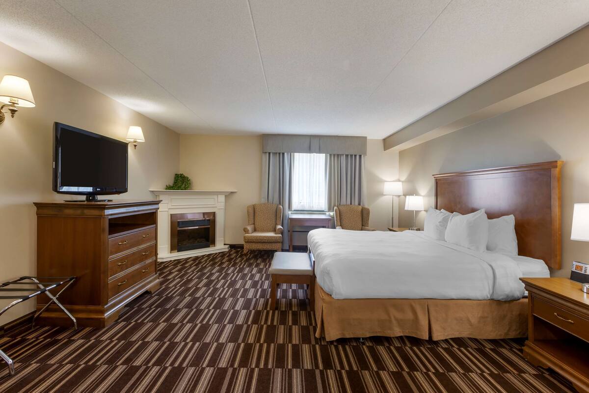 A hotel room with a large bed, a television, a desk with a chair, a fireplace, and a window with curtains.