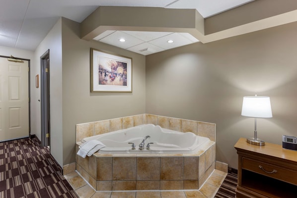 A hotel bathroom with a large bathtub, a framed picture on the wall, and a bedside lamp.