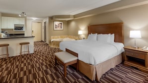 A hotel room with a large bed, a bench, a desk, a chair, a small table, a lamp, a phone, a microwave, a sink, and a picture on the wall.