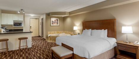 A hotel room with a large bed, a bench, a desk, a chair, a small table, a lamp, a phone, a microwave, a sink, and a picture on the wall.