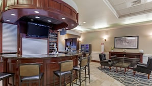 A bar area with a curved wooden counter, multiple bar stools, a television mounted above the bar, and a seating area with leather chairs and a coffee table.
