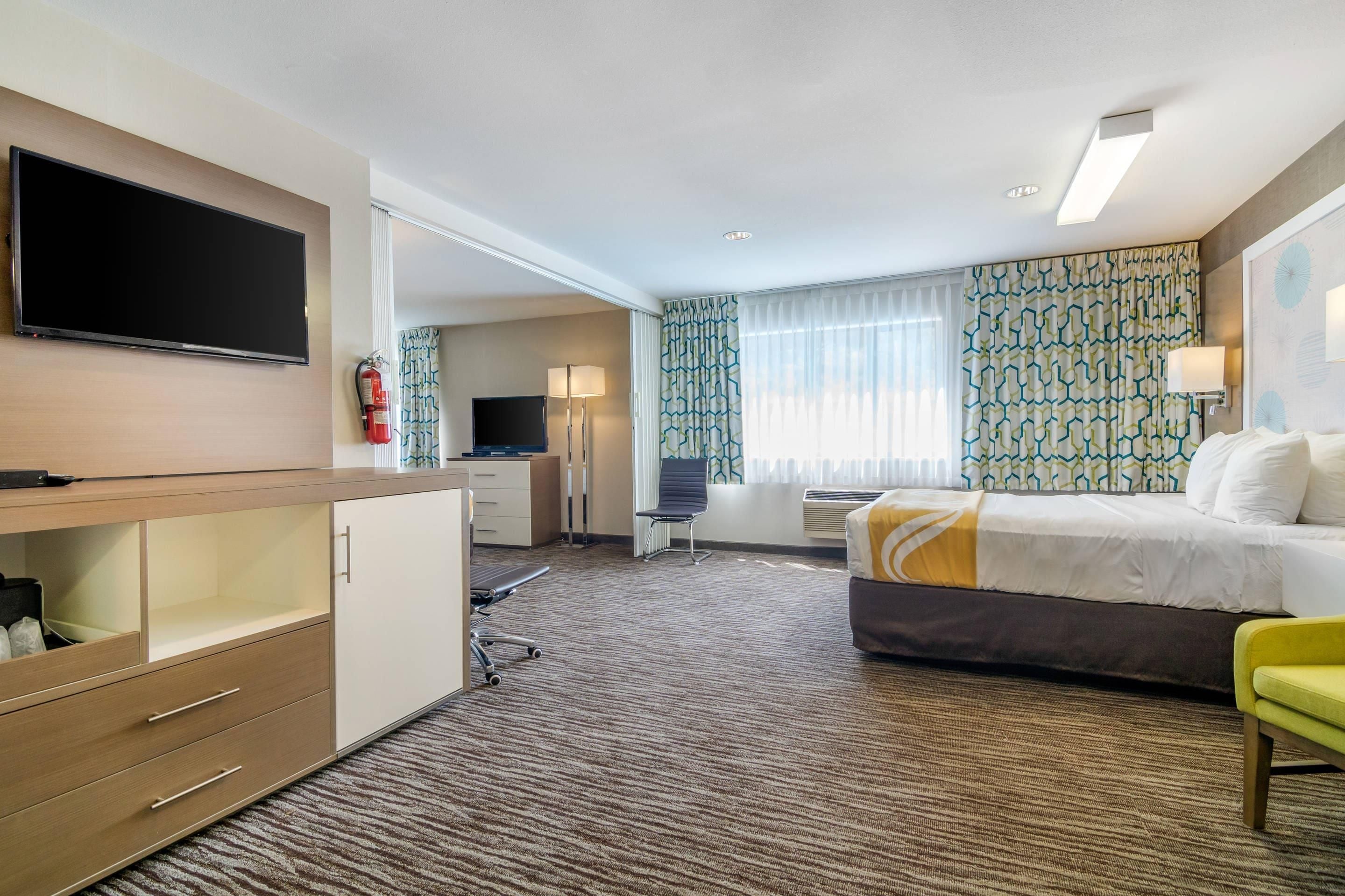 A hotel room with a bed, a desk, a chair, a television, and a fire extinguisher.