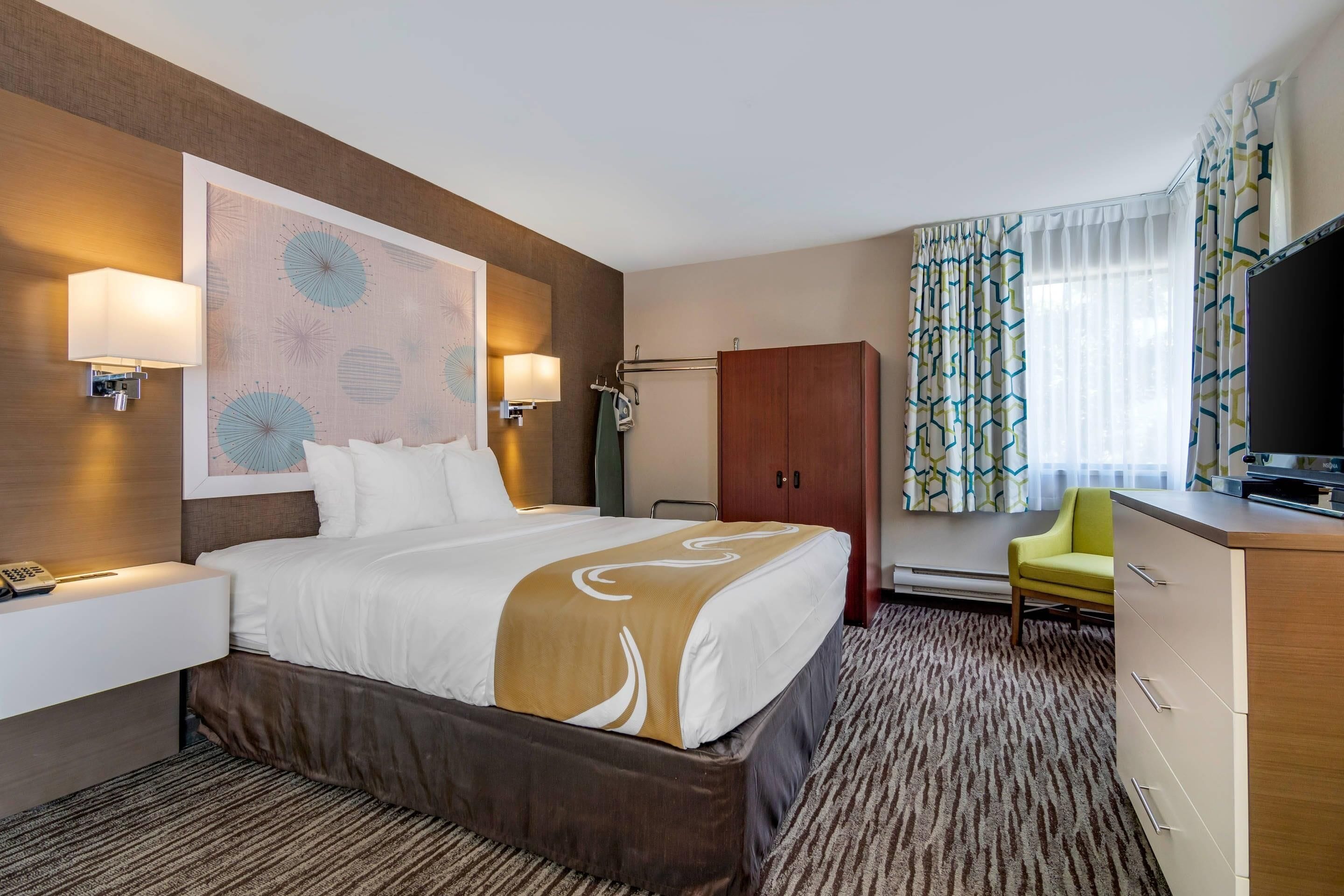 A hotel room with a large bed, a desk with a lamp, a television, and a wardrobe.