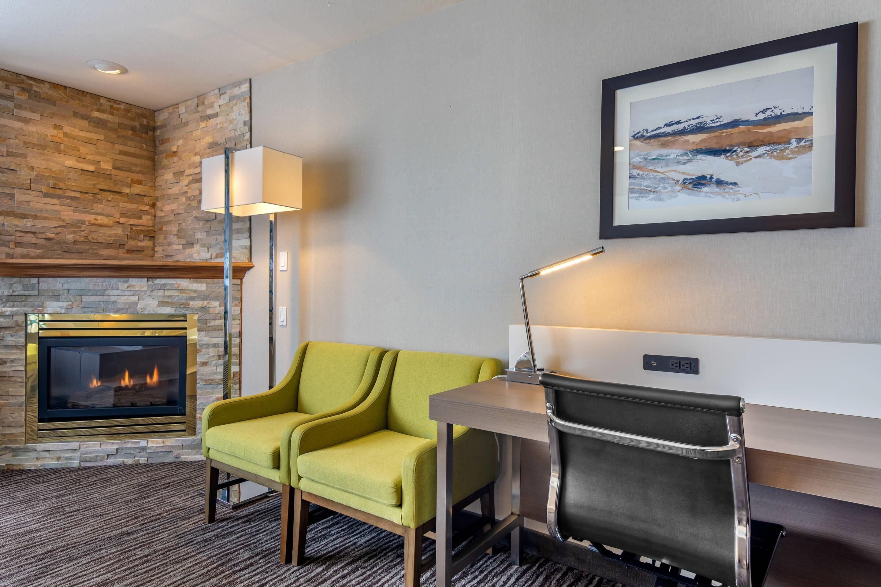 A modern hotel room with a stone fireplace, a green sofa, a desk with a chair, and a framed landscape painting on the wall.
