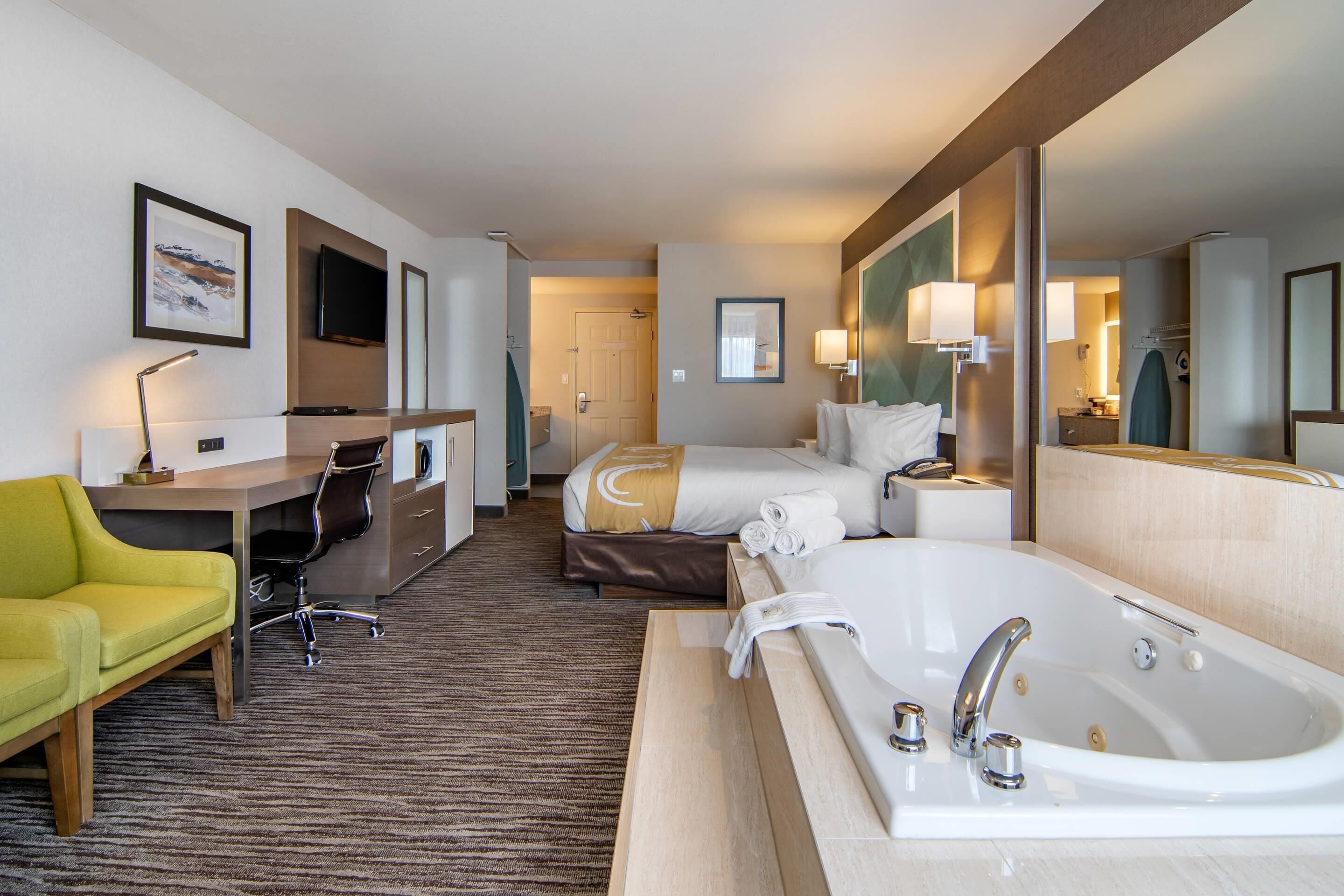 A hotel room with a bed, a desk, a chair, a large bathtub, and a television.