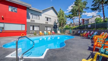 A swimming pool with a metal ladder, surrounded by colorful lounge chairs and a building with multiple windows.