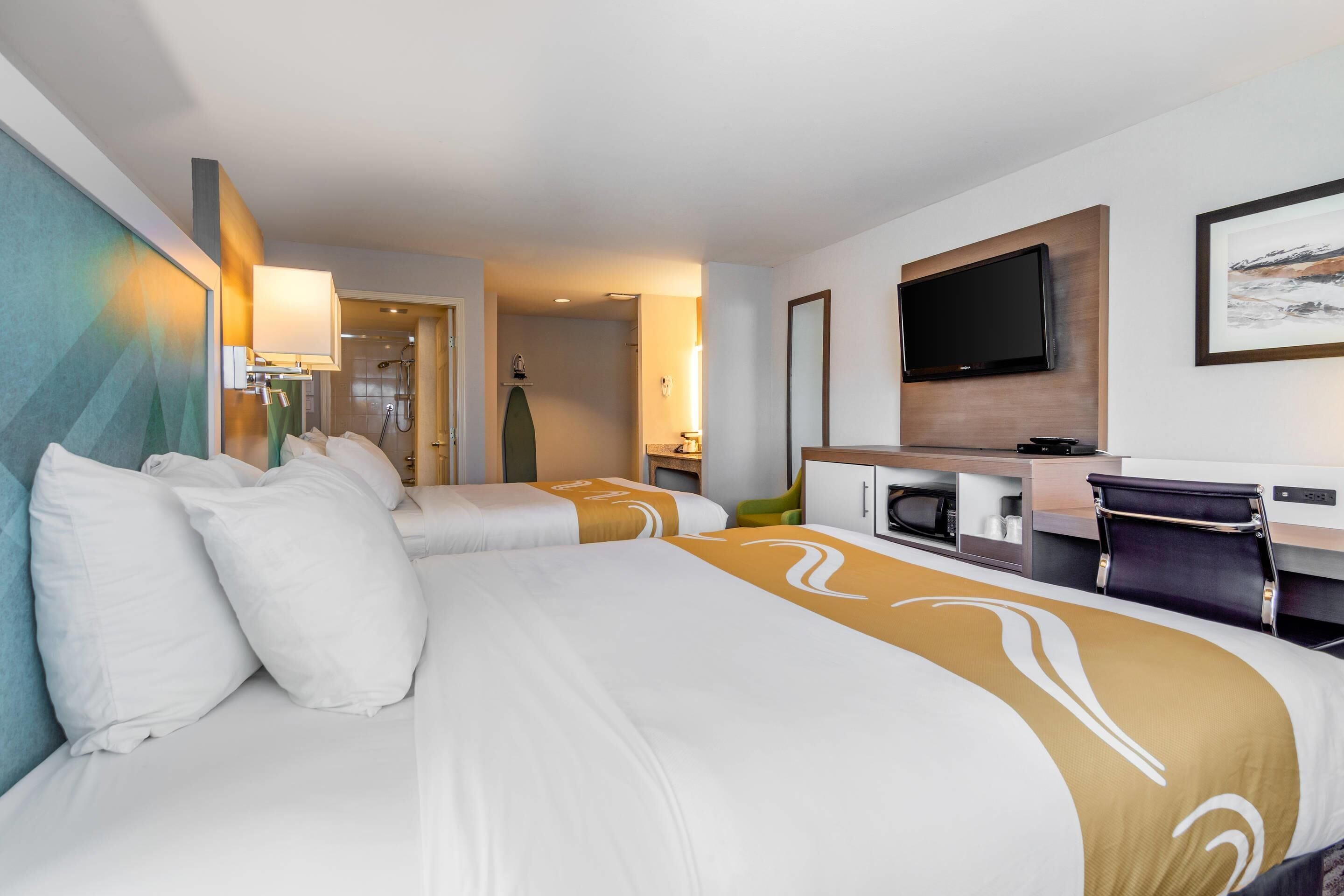 A hotel room with two beds, a desk, a TV, and a shower area.
