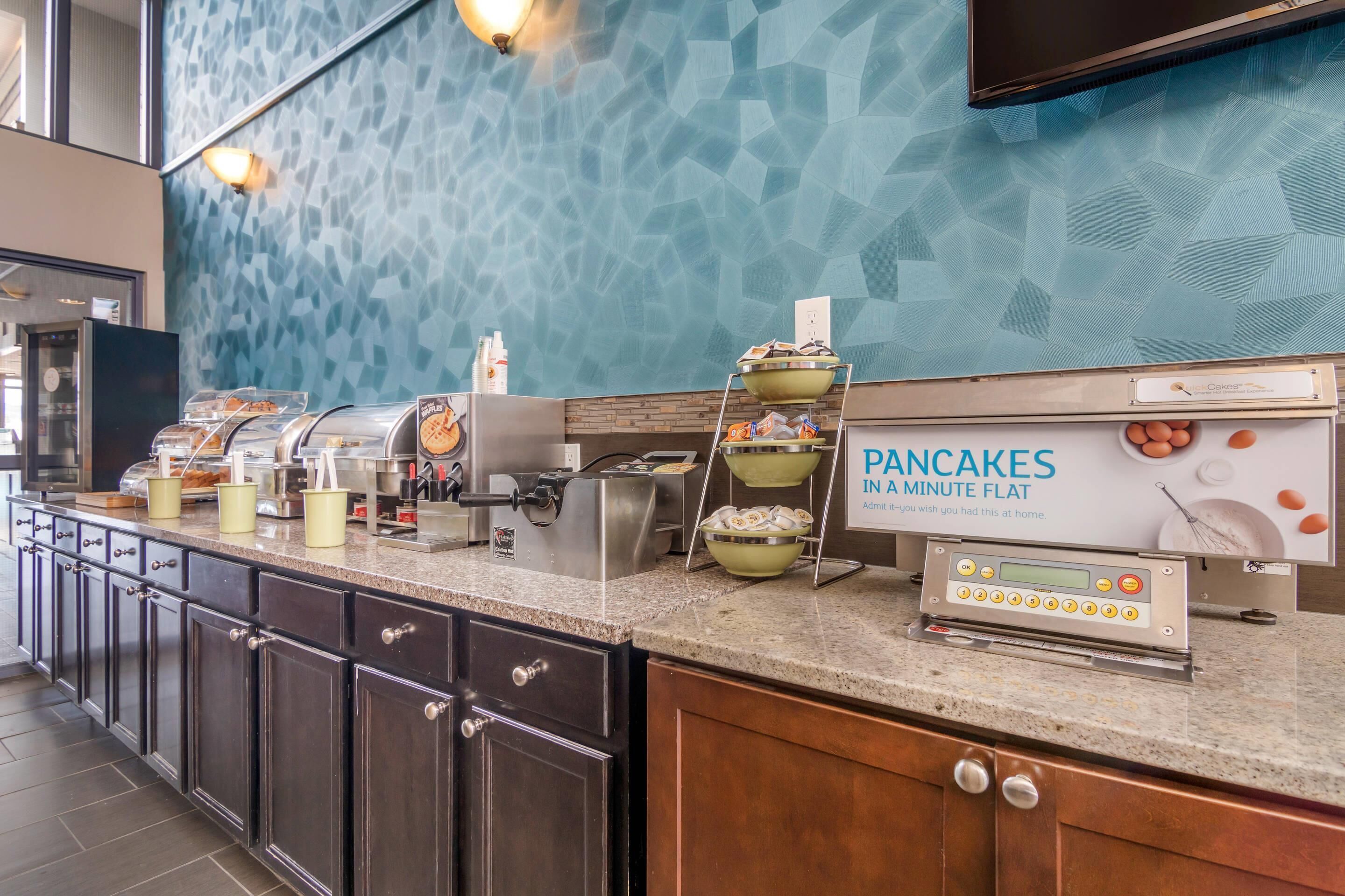 A breakfast station with a sign for pancakes, a coffee machine, and various food containers.