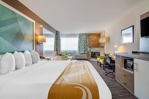 A hotel room with a large bed, a desk with a chair, a TV, a fireplace, and a view of the outdoors.