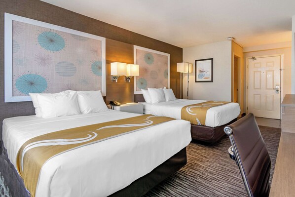 Two beds with white linens and gold accents, a large abstract art piece, and a desk with a chair and lamp.