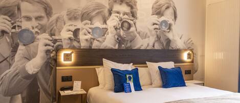 Habitación de hotel con una cama grande, un cabecero con luces, una mesita de noche con un teléfono y papeles, y una pared con una foto de personas.