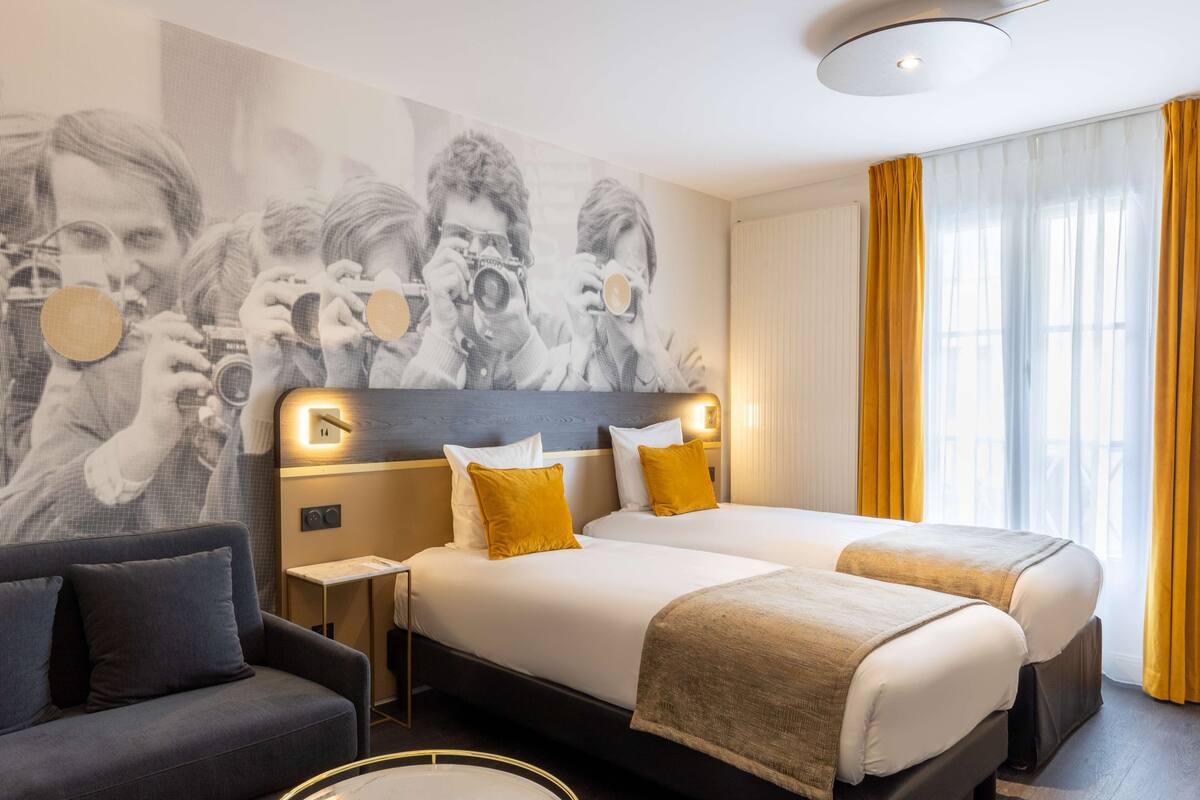 A hotel room with two beds, a sofa, a wall with a large photo, and a window with yellow curtains.