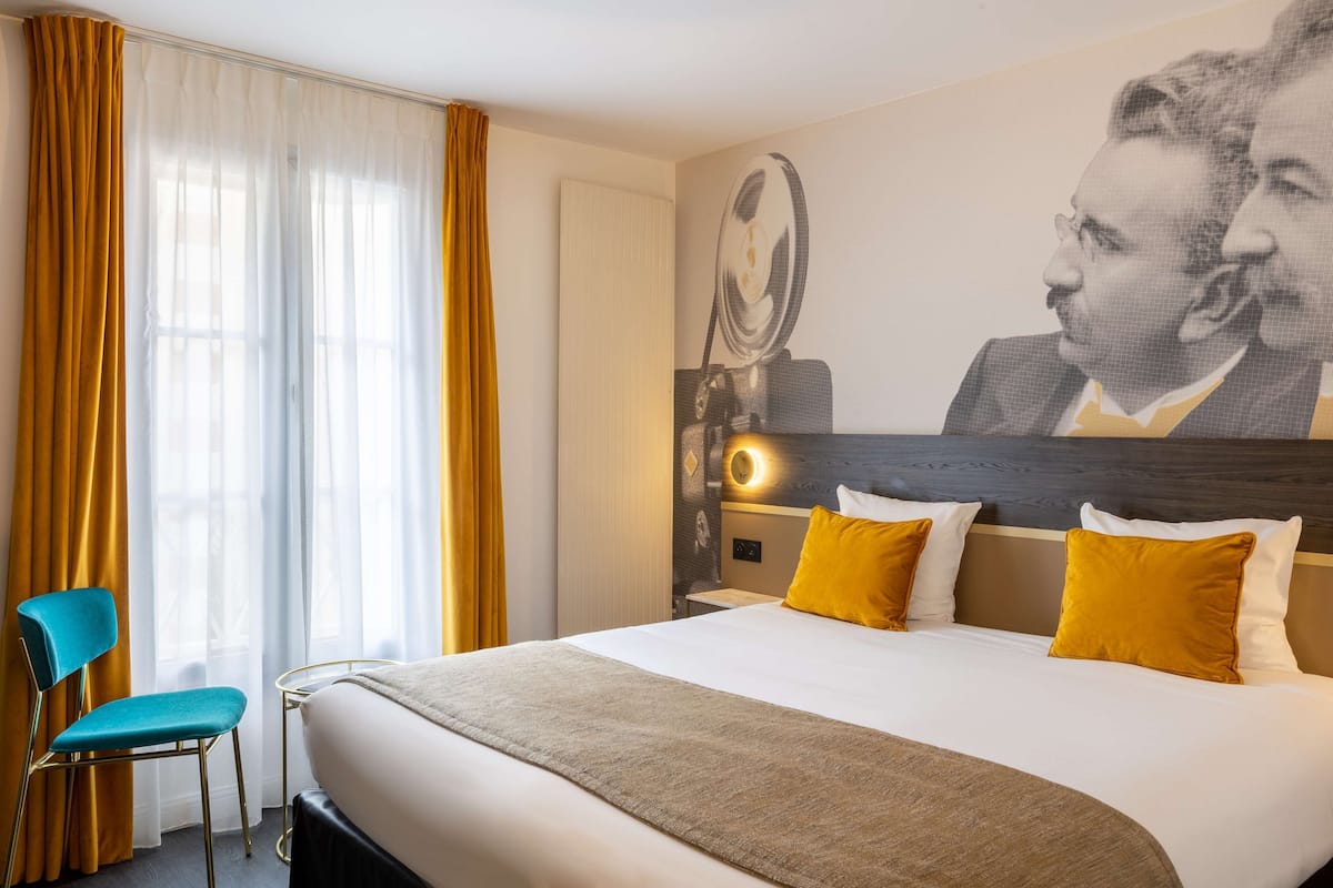 A hotel room with a large bed, a chair, a wall-mounted light, and a large portrait.