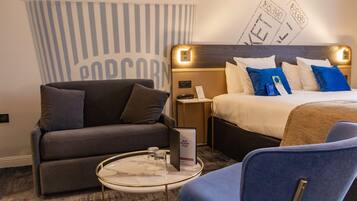 Quarto de hotel com cama, sofá, poltrona azul, pequena mesa redonda e decoração de parede com tema de pipoca.