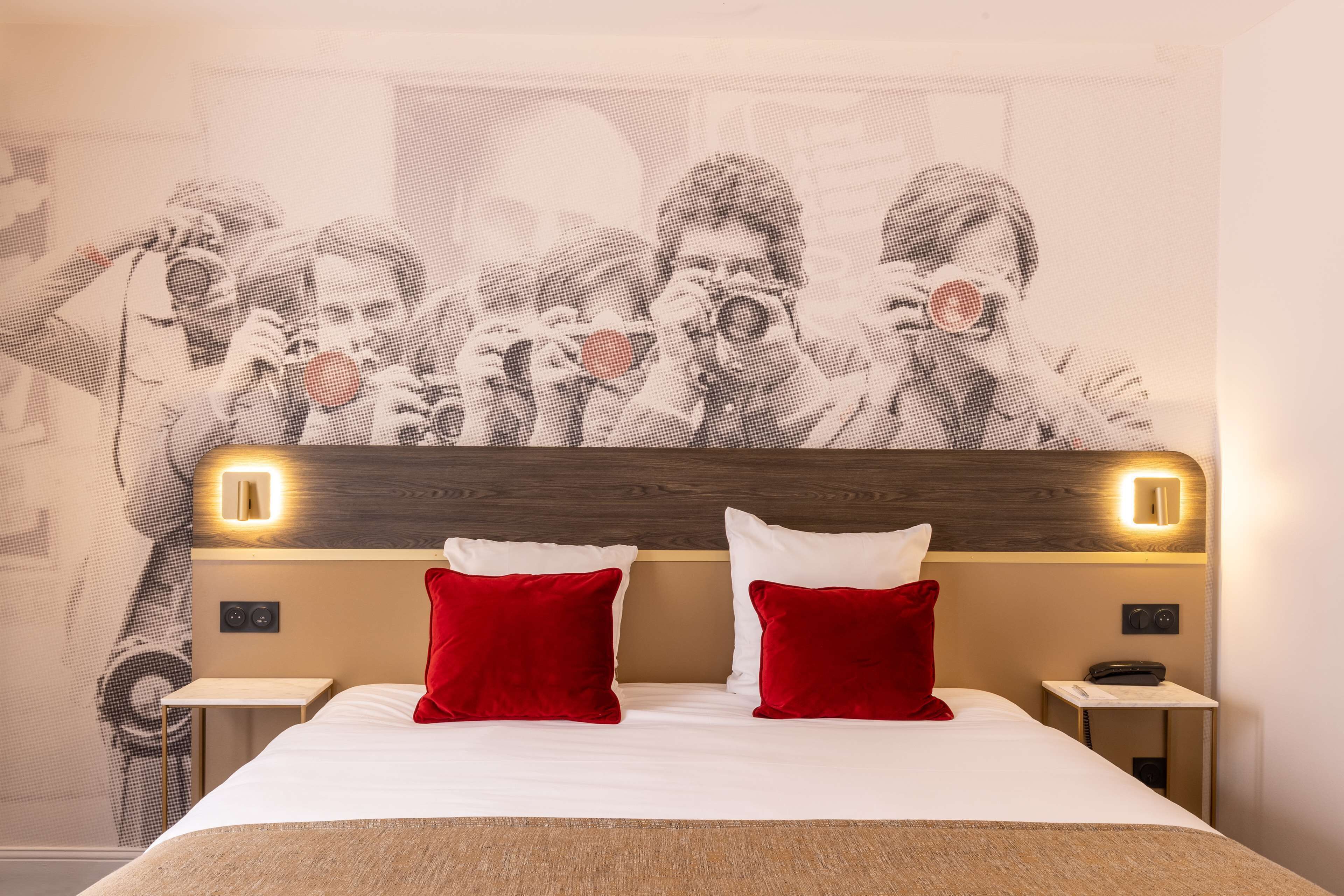 A hotel room with a large bed, red pillows, a headboard with integrated lighting, a nightstand with a phone, and a wall mural of people taking photos.