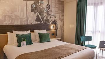 A hotel room with a large bed, green pillows, a headboard with a light, a green chair, and a wall with a vintage photo.