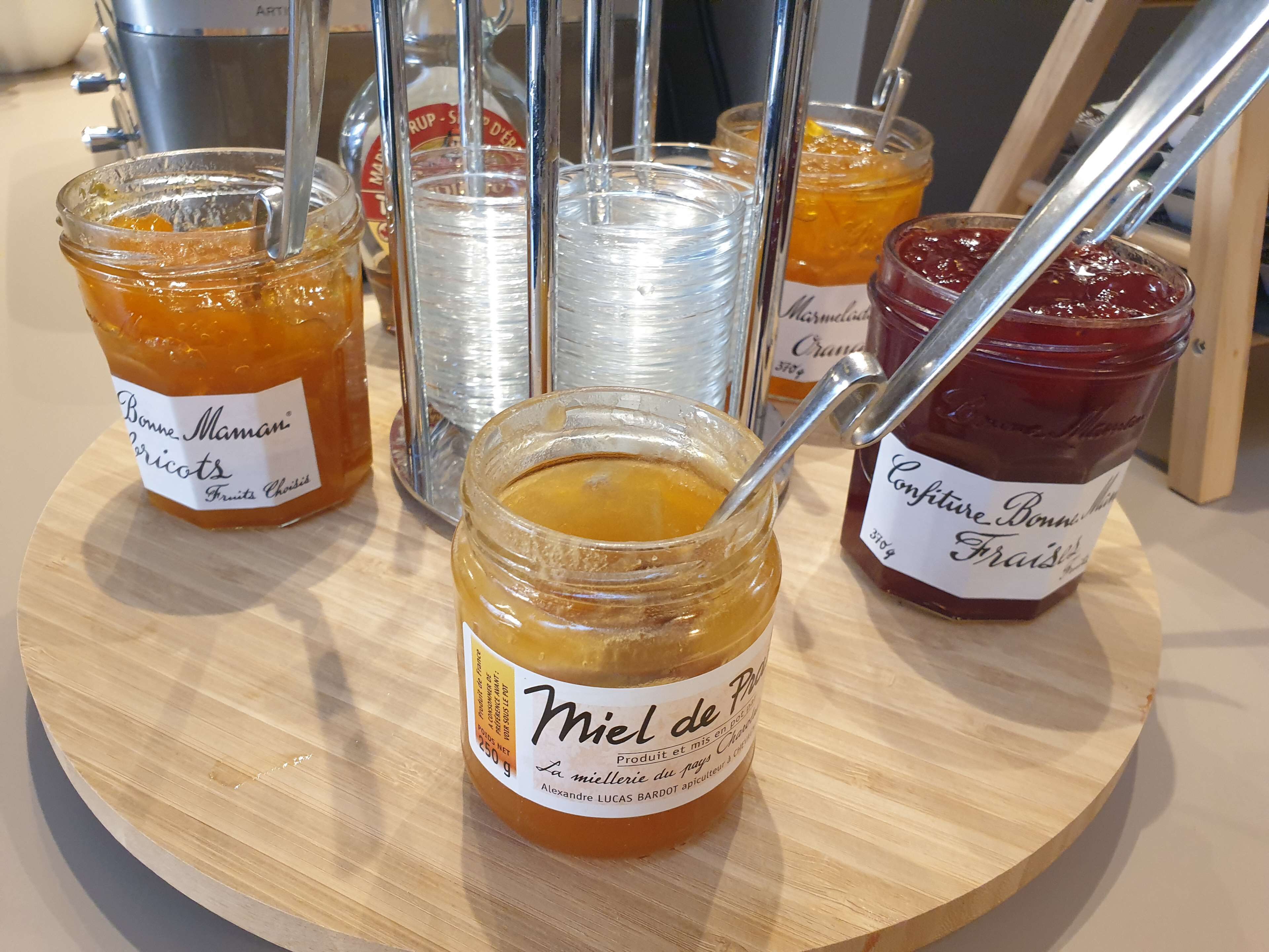 Une planche en bois sur laquelle sont disposés différents pots de miel et de confiture, ainsi que des étiquettes et des cuillères.